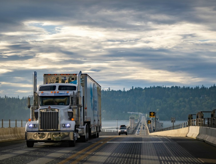 A white semi-truck driving across a long bridge over water, with forested hills and a cloudy sky in the background.