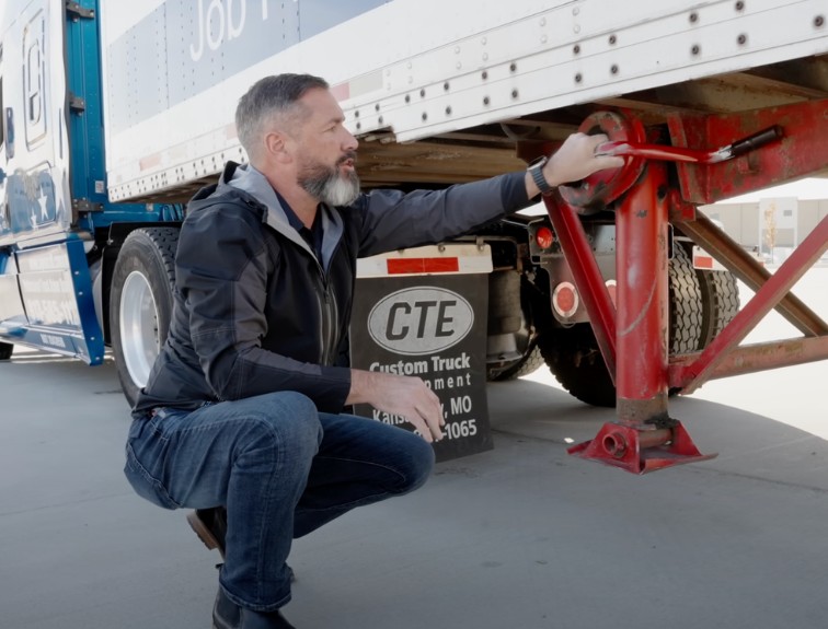 A man crouching beside a semi-trailer during a pre-trip inspection, checking the red landing gear next to a CTE Custom Truck Equipment placard.
