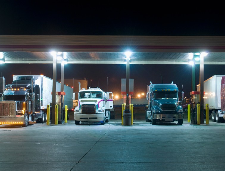 Semi-trucks parked at fuel islands under a brightly lit truck stop canopy at night.