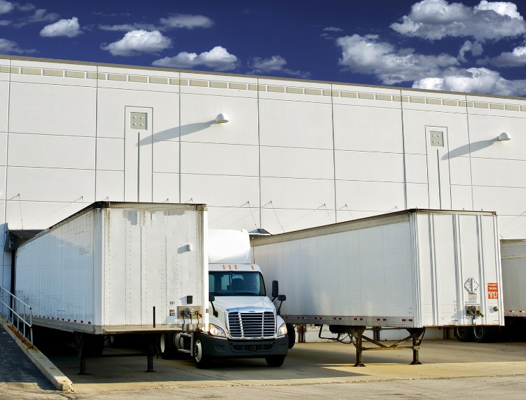 A white semi-truck parked between two trailers at a warehouse loading dock under a blue sky with scattered clouds.