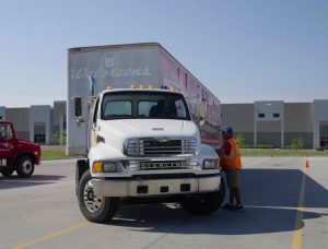 A trainer in an orange safety vest talks with a student driver through the window of a white truck pulling a Walgreens trailer on a training lot