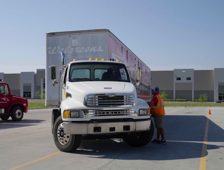 A trainer in an orange safety vest talks with a student driver through the window of a white truck pulling a Walgreens trailer on a training lot